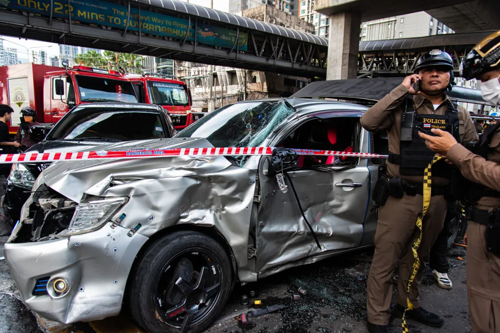 A heavily damaged silver car at an accident scene with police tape, police officers, and a fire truck in the background.