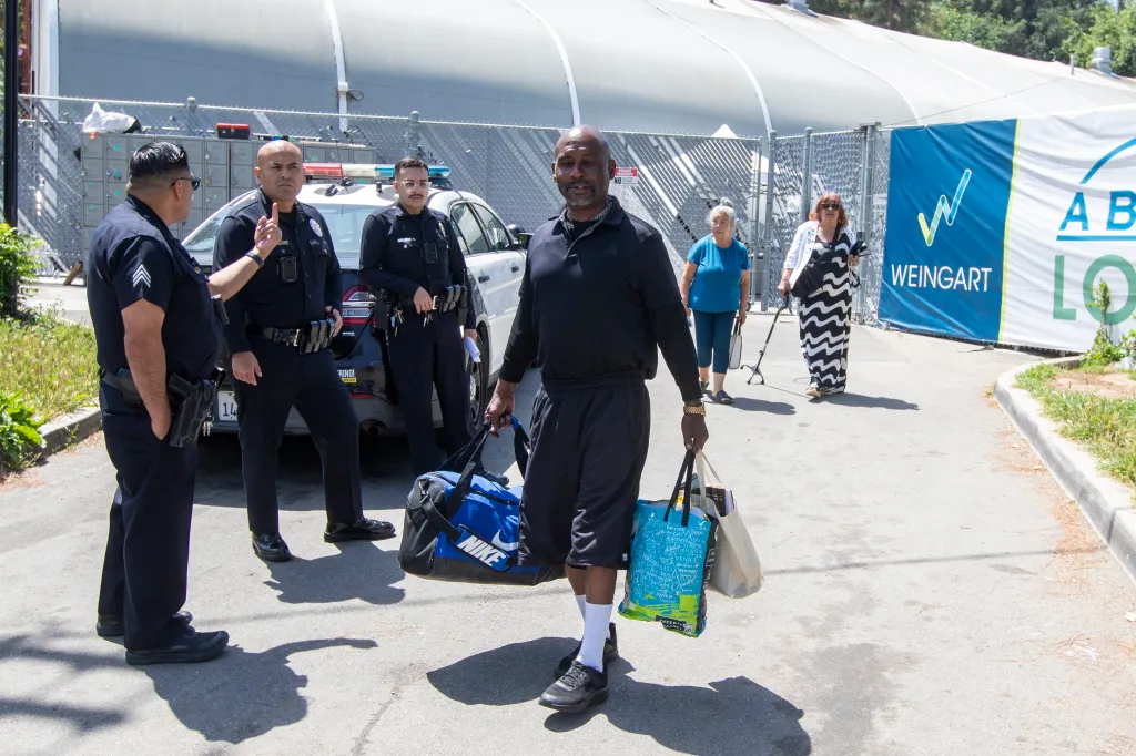 Police officers watch a man carrying his belongings leave a homeless shelter.