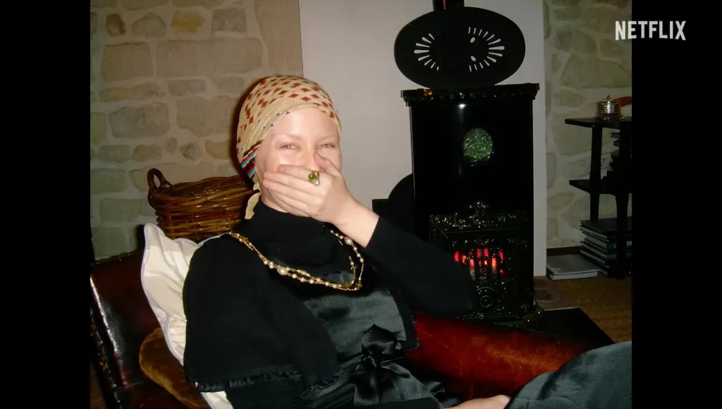 A woman with a patterned headscarf and a black dress covering her mouth with her hand, sitting in a room with a fireplace.