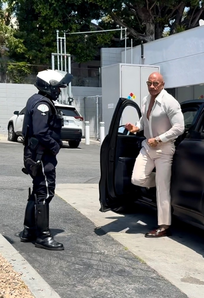 Dwayne Johnson in a white shirt and pants gets pulled over by a motorcycle police officer for tinted windows in Hollywood.