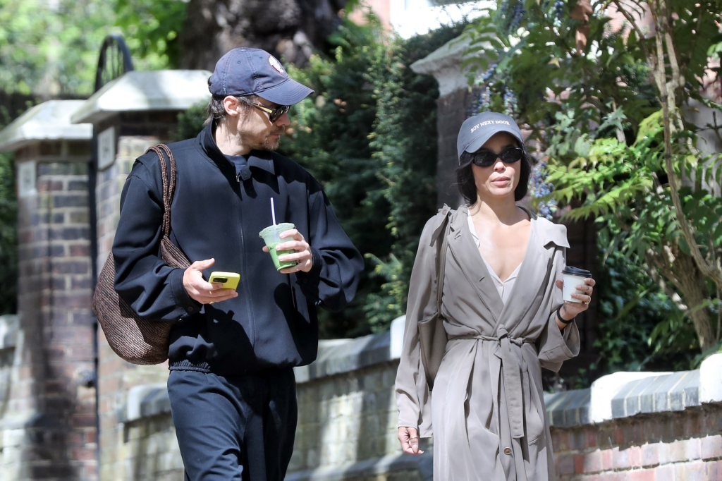 Harry Styles in a navy jacket and Zoe Kravitz in a beige trench coat walk down a street holding drinks.