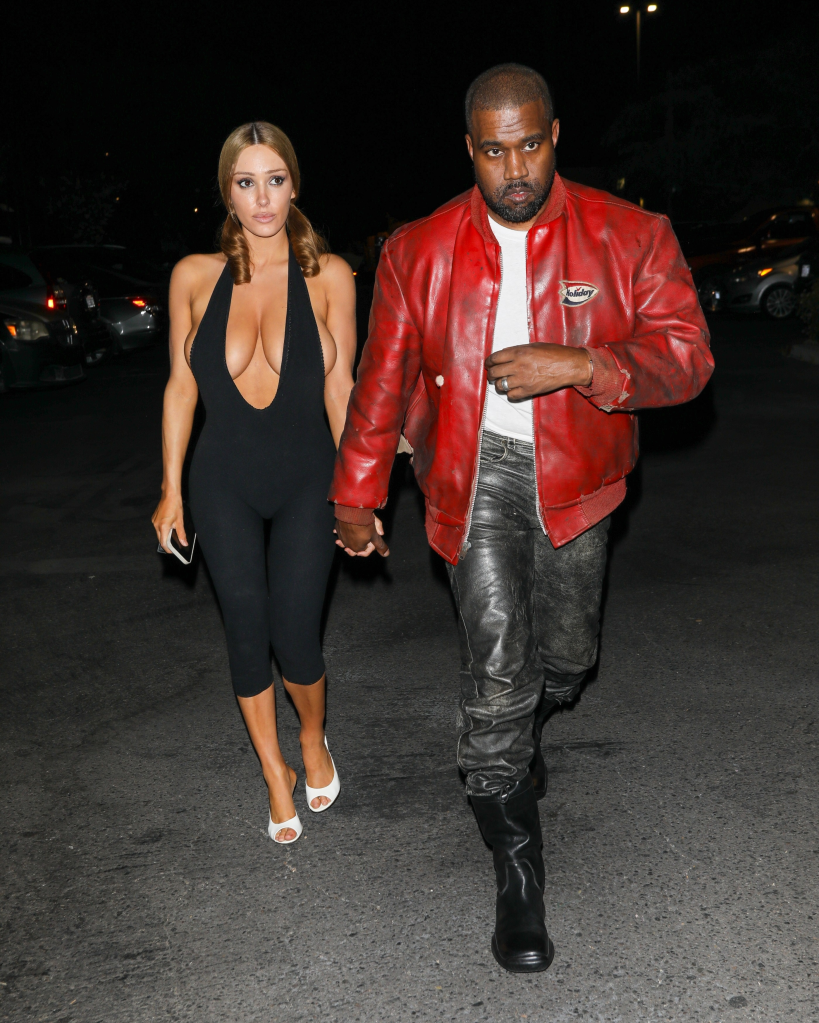 Kanye West in a red jacket and Bianca Censori in a black halter jumpsuit walk together.