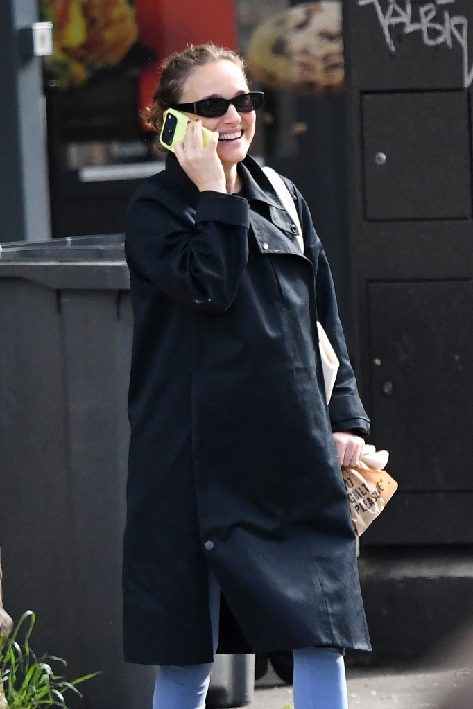 Natalie Portman, pregnant, talks on a neon green phone while wearing a black coat, sunglasses, blue leggings, and white sneakers, carrying a beige tote bag and a paper takeaway bag.