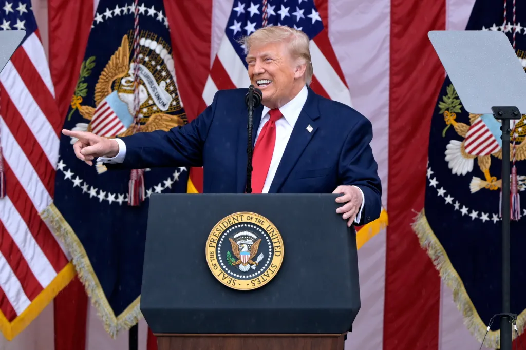 President Donald Trump speaking at a podium with the Seal of the President of the United States, against a backdrop of US flags and presidential flags.