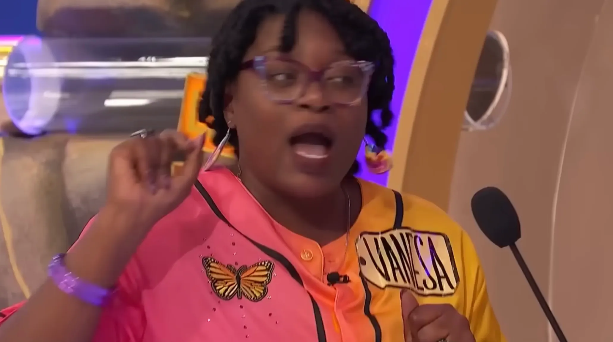 A woman named Vanessa in a split pink and yellow shirt with a butterfly design, speaking into a microphone.