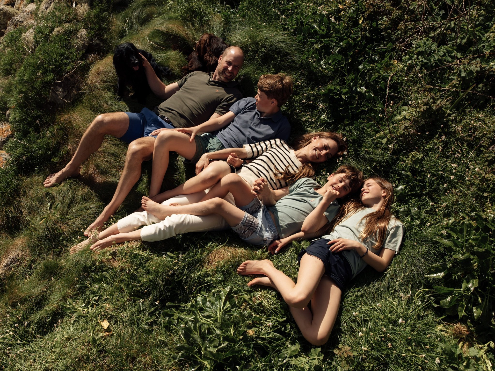 The Prince and Princess of Wales with their children and dog lying in grass.