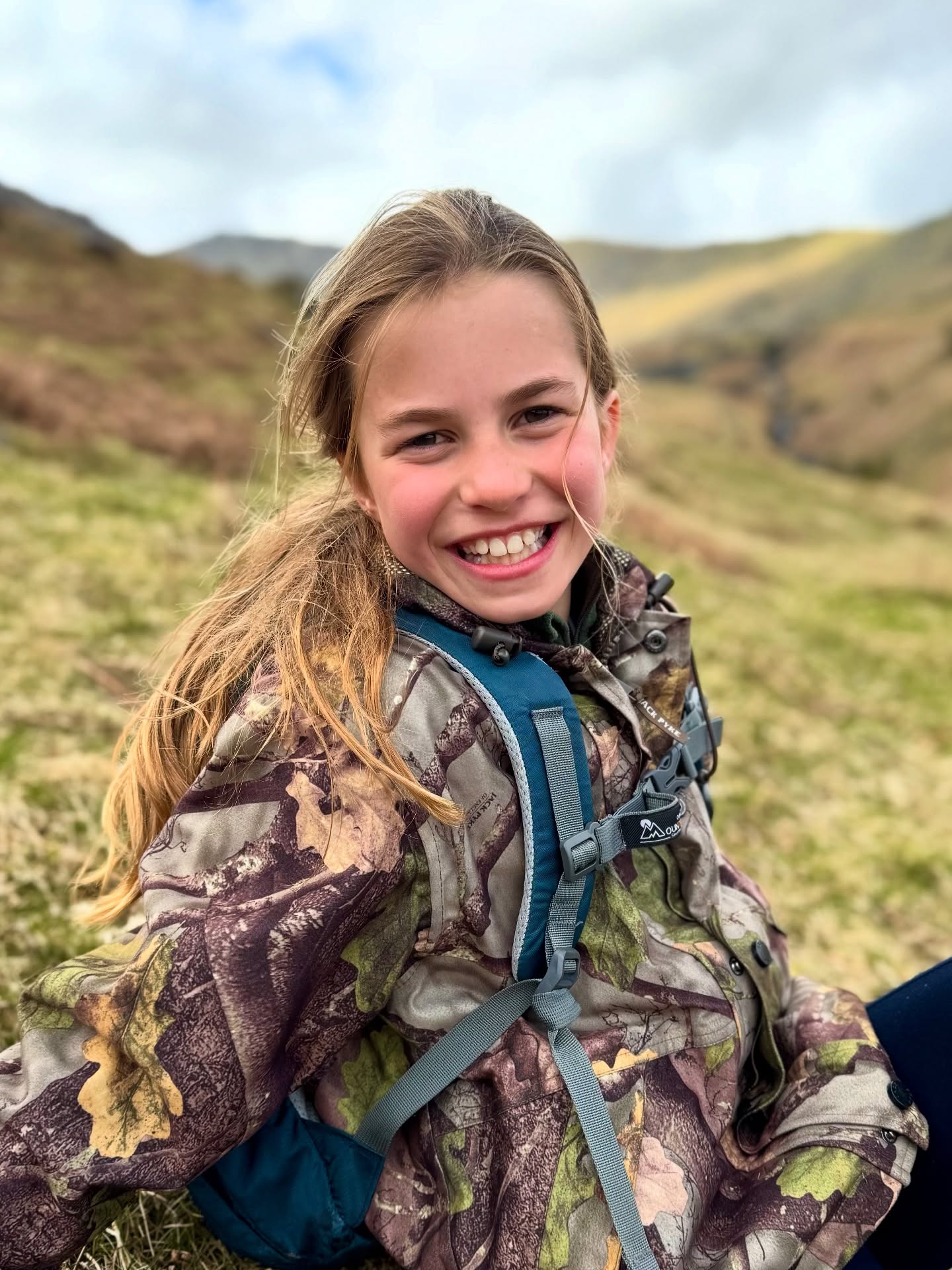 Princess Charlotte in a camouflage jacket and backpack, smiling in a mountainous, grassy landscape.