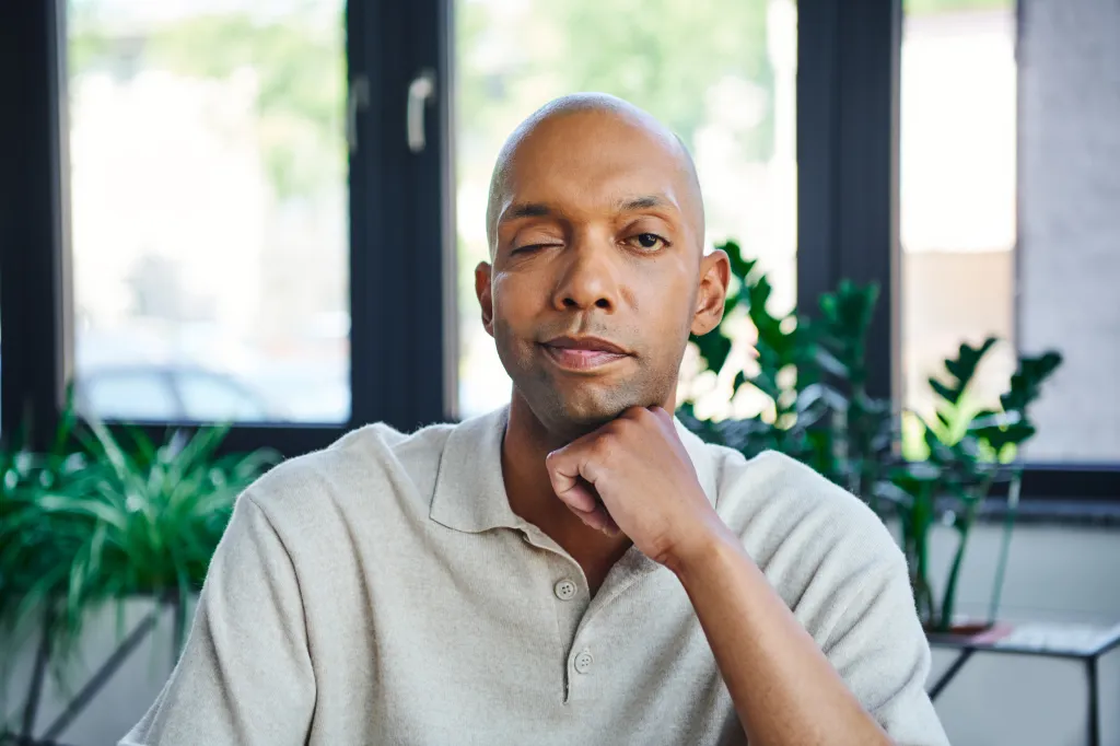 A Black man with ptosis and a shaved head, wearing a cream polo shirt, gazes thoughtfully at the camera with one eye.