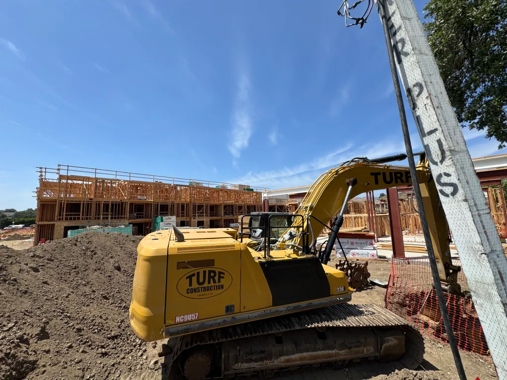 Construction site with a yellow excavator from Turf Construction in the foreground and the wood frames of new housing units in the background.