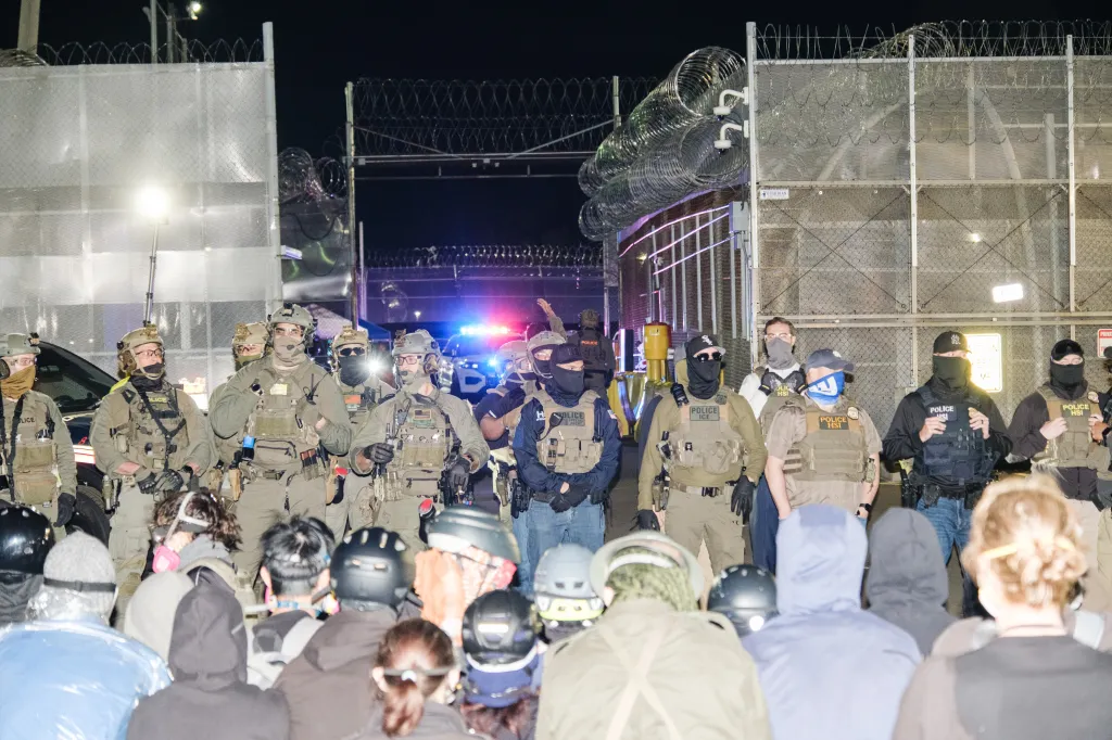 Protesters facing a line of ICE officers in tactical gear at night outside a detention facility.