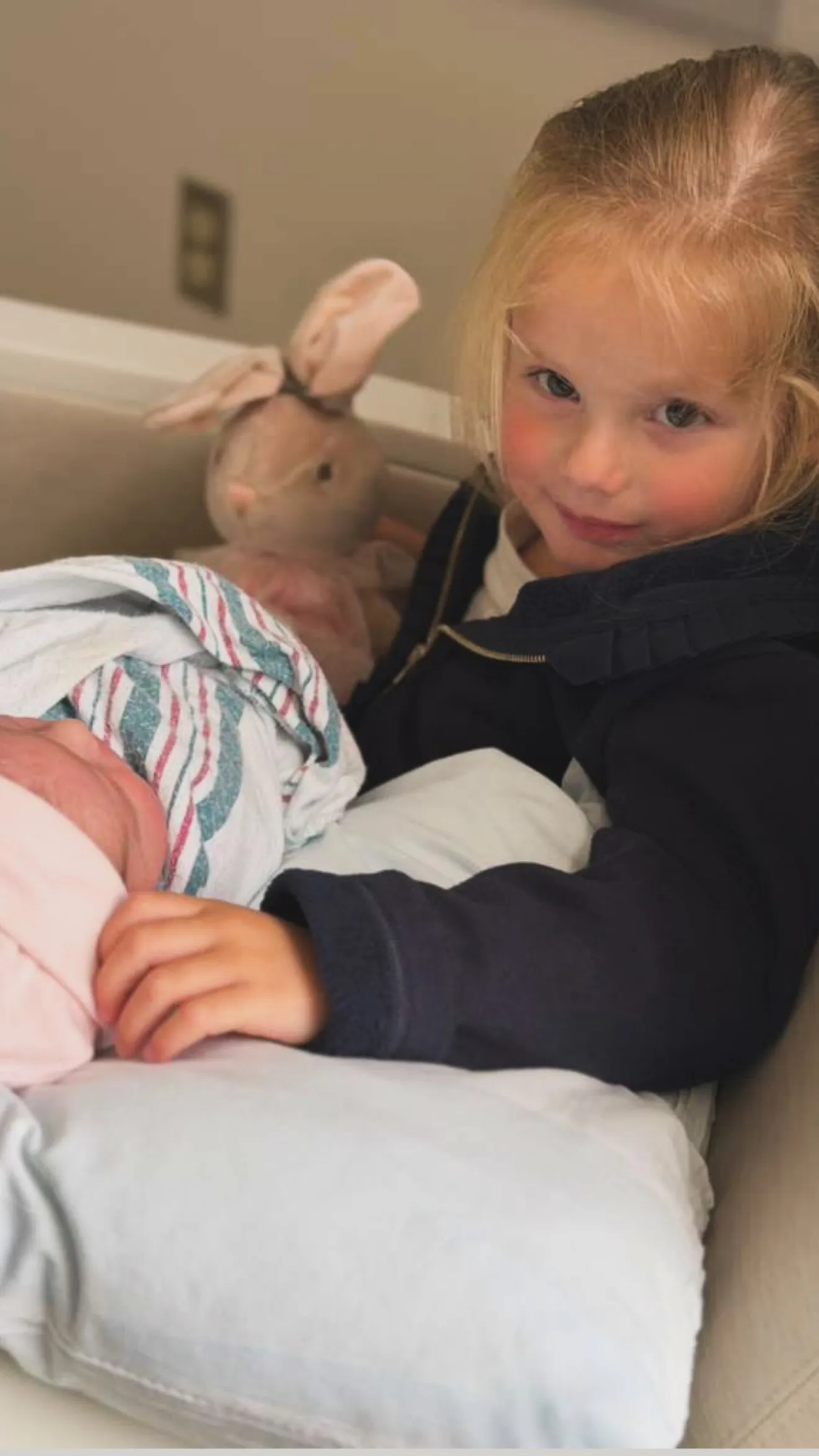Royce, a young child, is seated next to a swaddled infant (Rose) and a stuffed bunny, smiling at the camera.