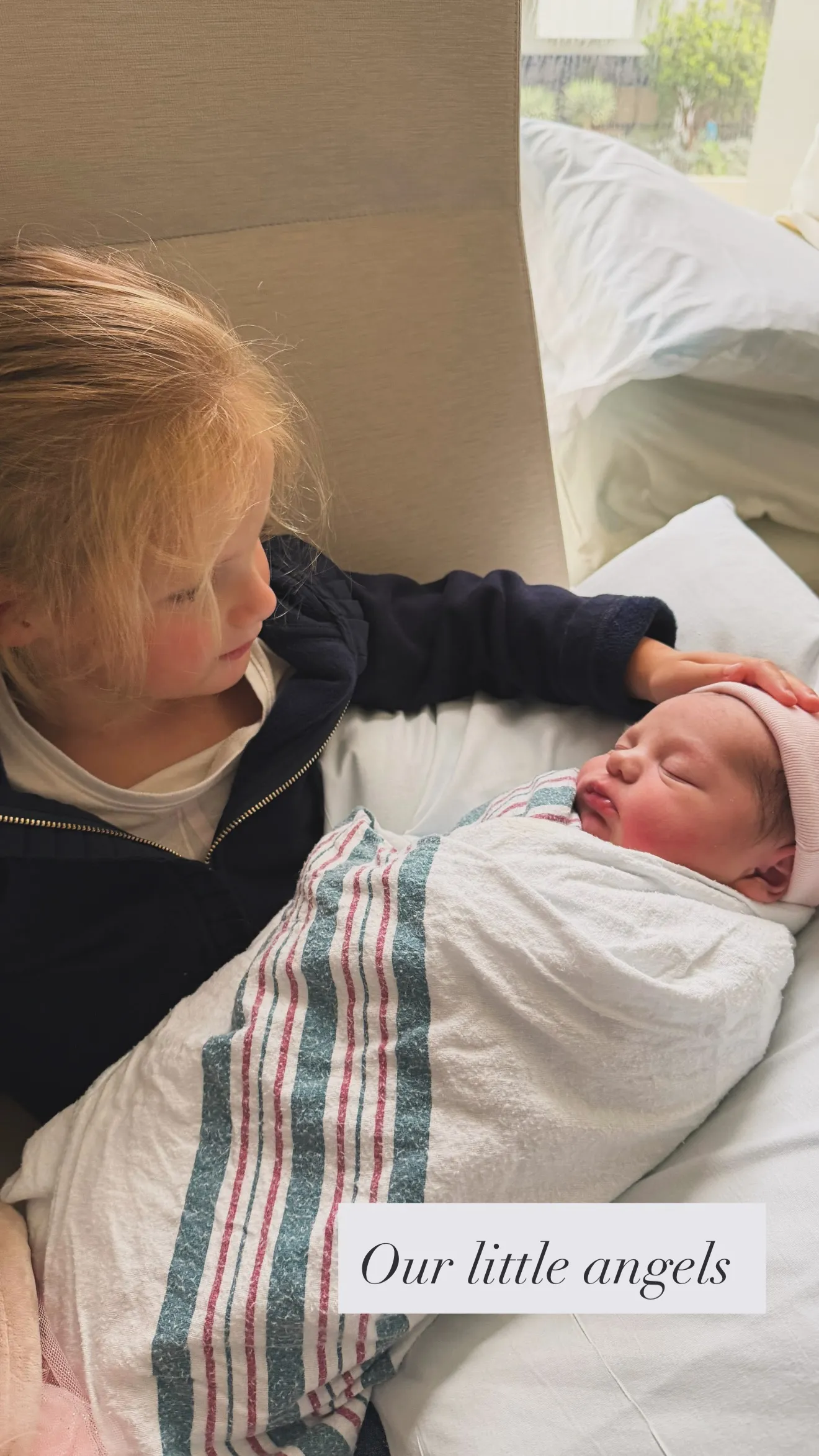 A young girl gently touching a newborn baby wrapped in a striped blanket.
