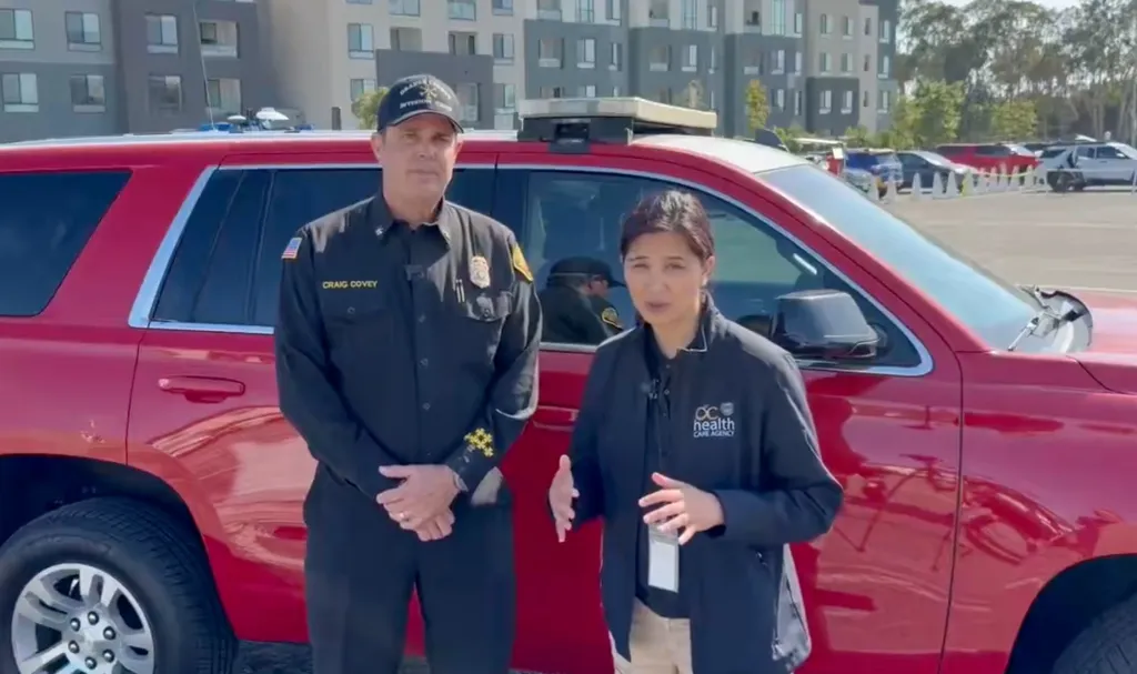 OCFA Division Chief Craig Covey and Dr. Regina Chinsio-Kwong of Public Health stand next to a red SUV, warning about health problems from toxic chemical leaks.