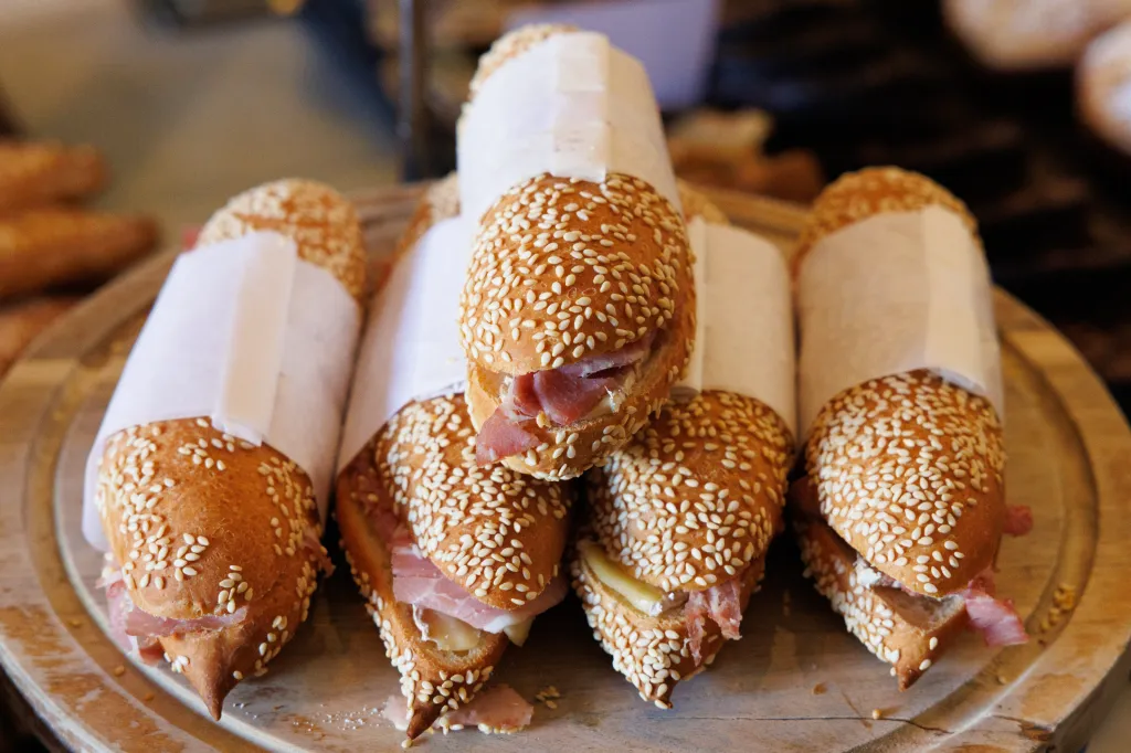 Small baguette sandwiches with sesame seeds and prosciutto, wrapped in white paper.