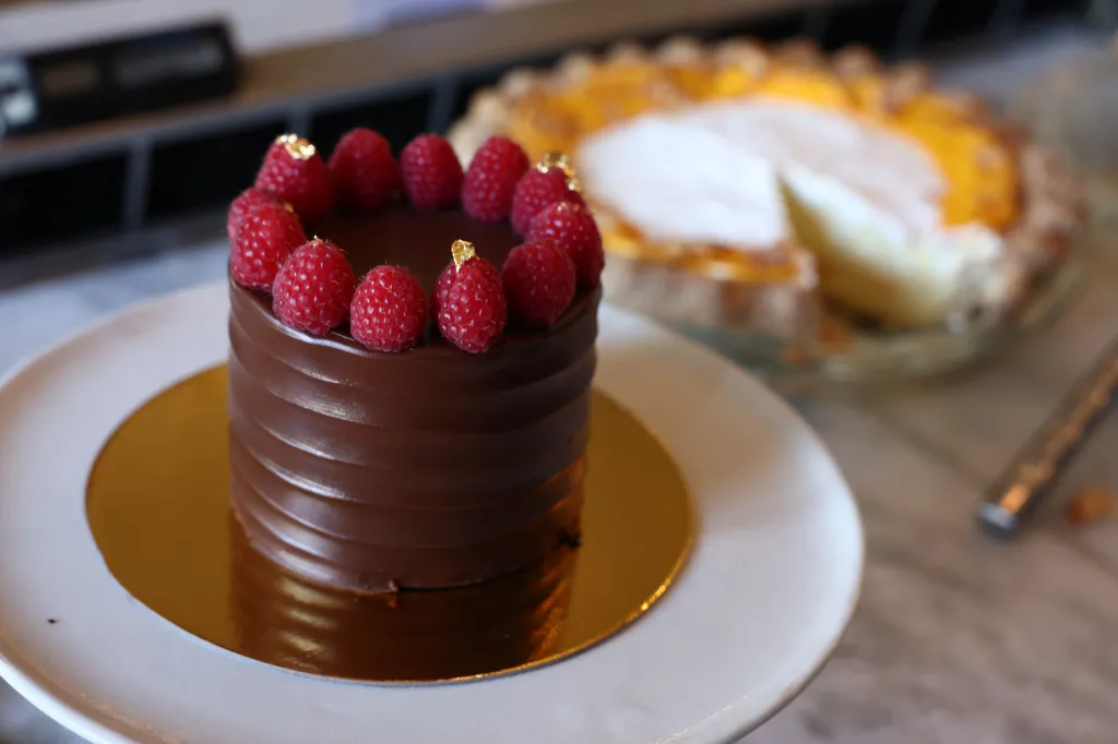 A chocolate cake topped with raspberries and gold flakes, with a tart in the background.