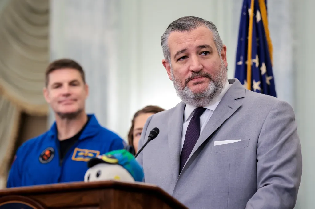 Senator Ted Cruz speaks at a podium with an Artemis II crew member in the background.