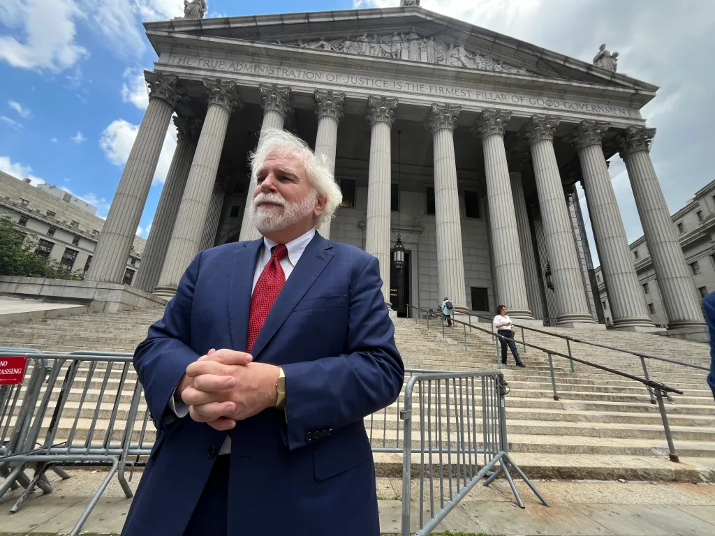 Randy Mastro in a blue suit and red tie stands in front of a classical building with pillars, his hands clasped.