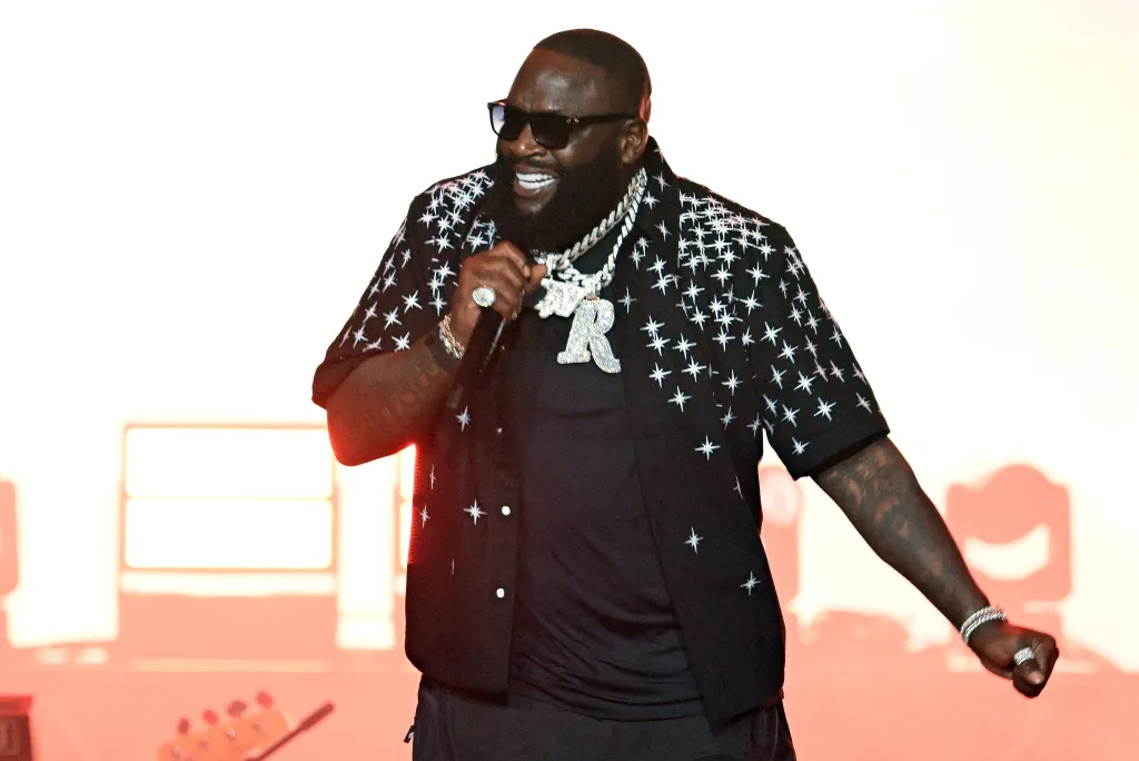 Rapper Rick Ross performs onstage in a black shirt with white starburst designs, wearing sunglasses, and two diamond-encrusted necklaces.