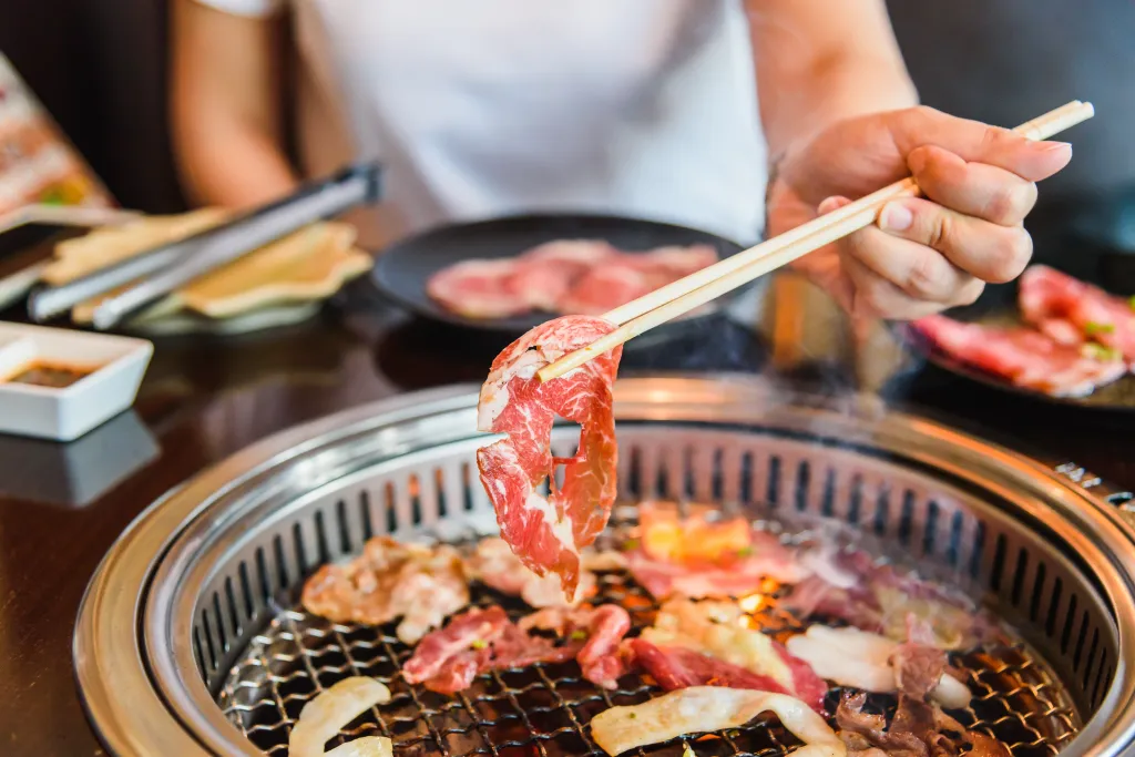 A hand holding a thin slice of raw marbled beef with chopsticks over a yakiniku grill.