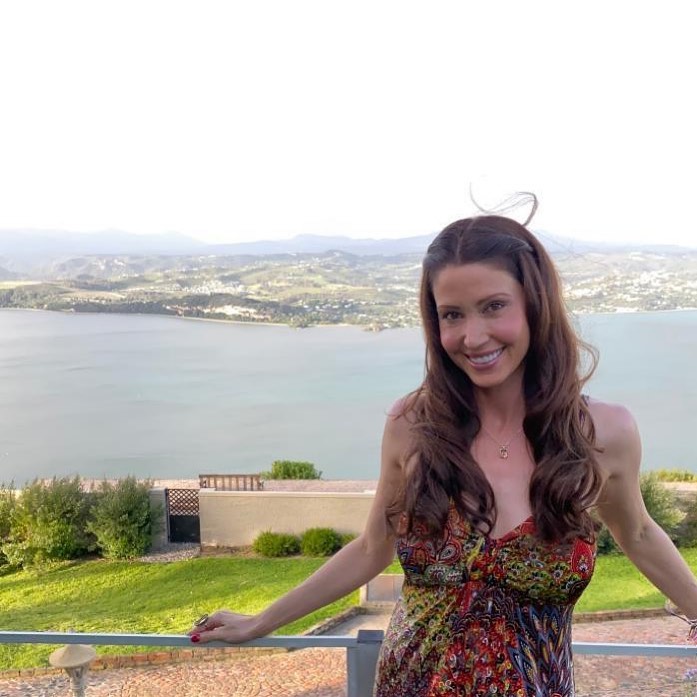 A woman with long brown hair, wearing a colorful patterned dress, smiles while leaning on a railing with a large body of water and hills in the background.