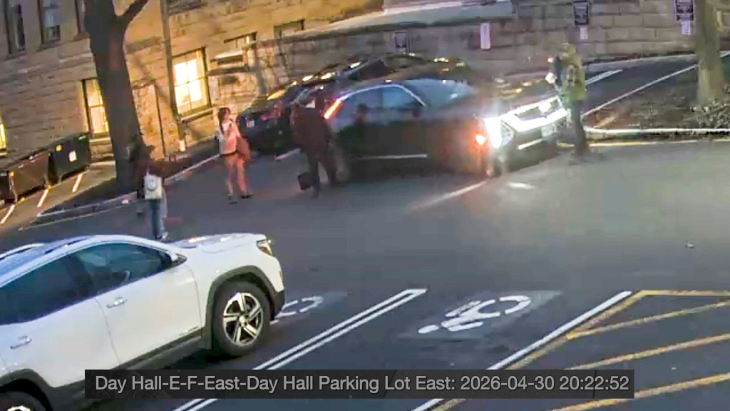 Still image from a video showing several people in a parking lot, with one person next to a black car with its lights on.