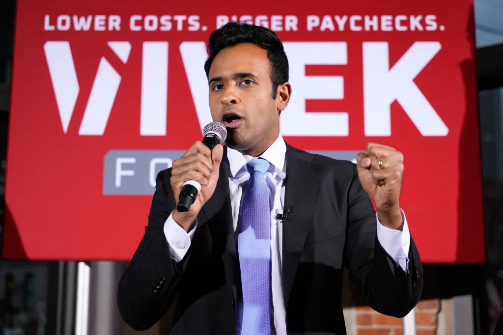 Vivek Ramaswamy speaking into a microphone with a clenched fist, in front of a red banner that reads 