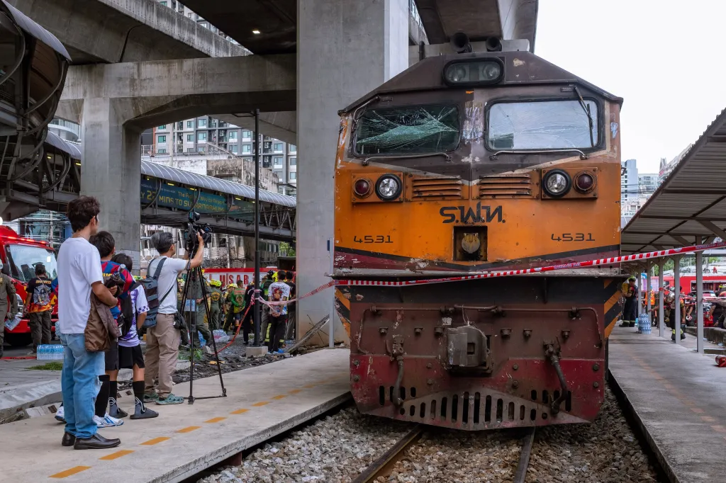 A train with a shattered front window is on the tracks next to a platform, cordoned off by red and white tape, with emergency services and news crews in the background after a train-bus collision in Bangkok.
