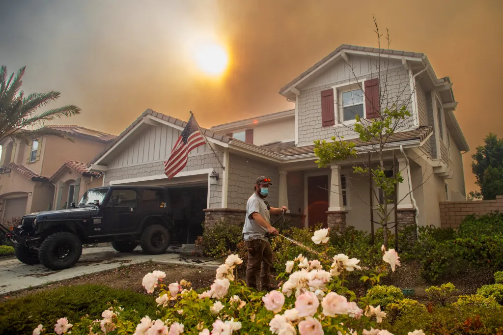 A resident waters plants with a hose outside a house under a smoky sky as the Sandy Fire burns in the hills in Simi Valley, California.