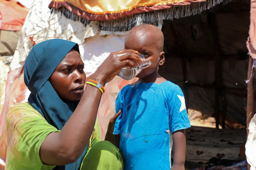 A woman in a teal hijab and green top helps a small child in a blue shirt drink from a glass.