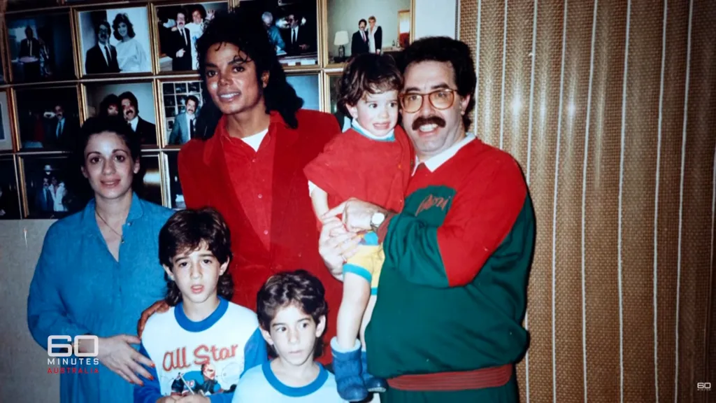 Michael Jackson posing with the Cascio family, a woman, a man holding a young boy, and two other young boys in front of a wall of framed photos.