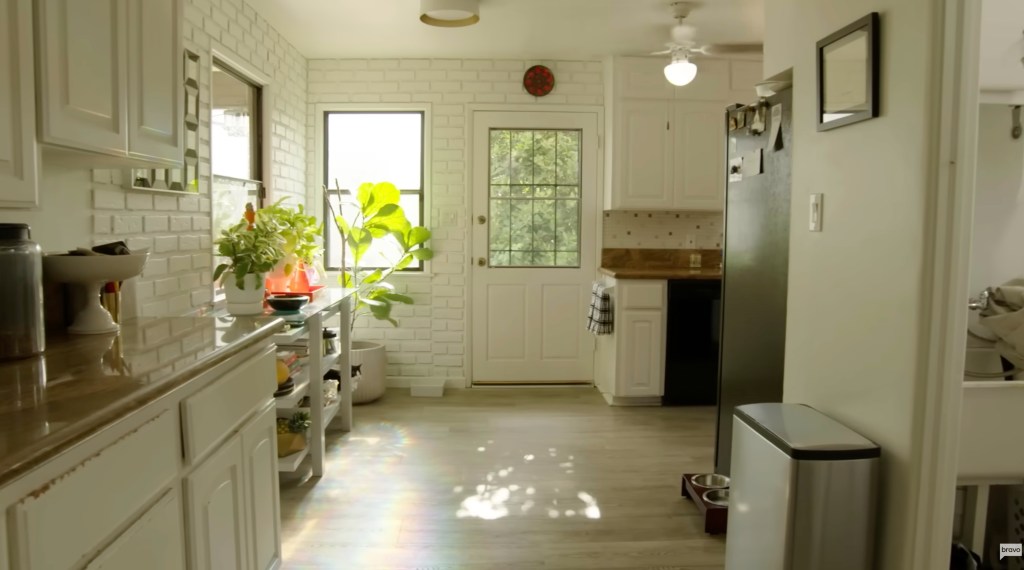 A modern kitchen with white brick walls, white cabinets, and light wood flooring.