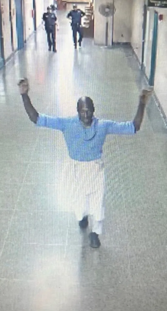 Surveillance photo of Rhamell Burke with his arms raised, as two officers approach him from behind.