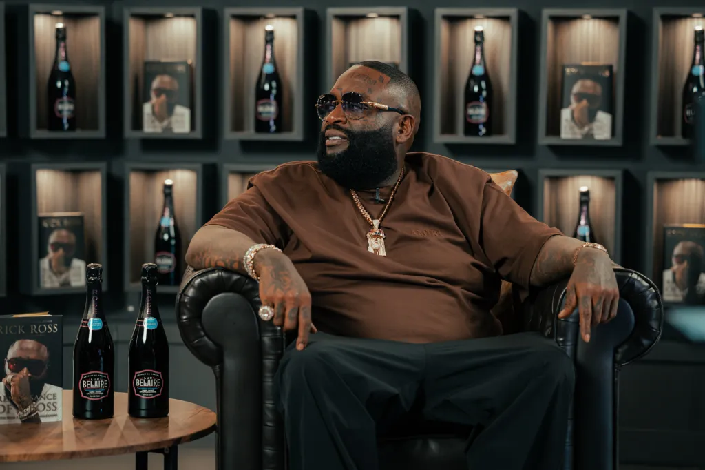 Rick Ross sitting in a black leather chair with bottles of Luc Belaire and a book titled 