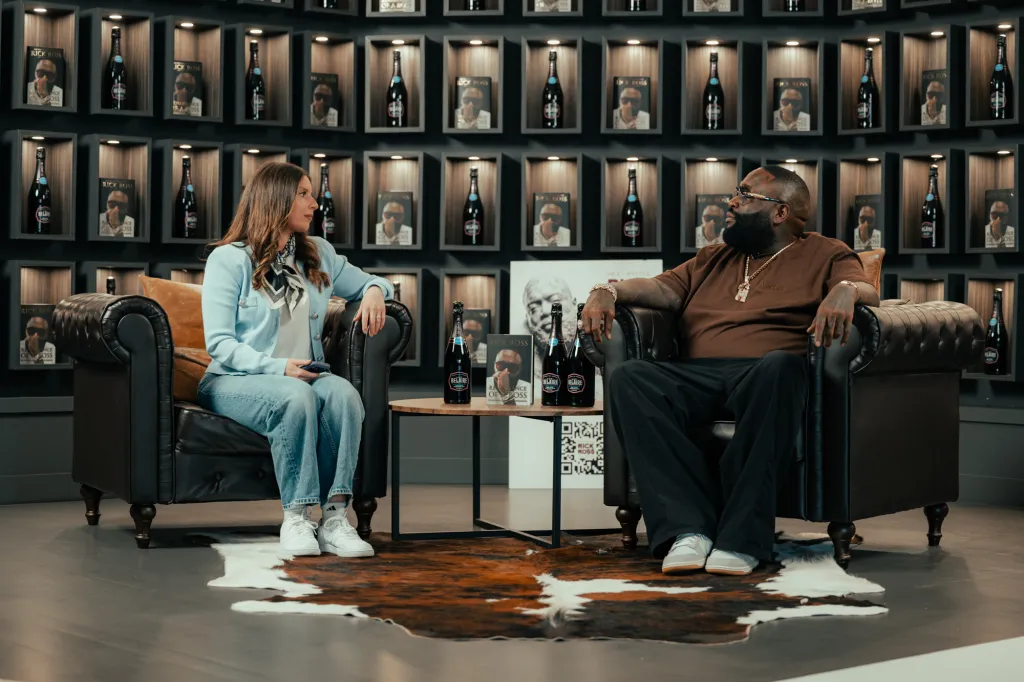 Rick Ross being interviewed by a woman, sitting in leather armchairs in a room with shelves displaying bottles and books.