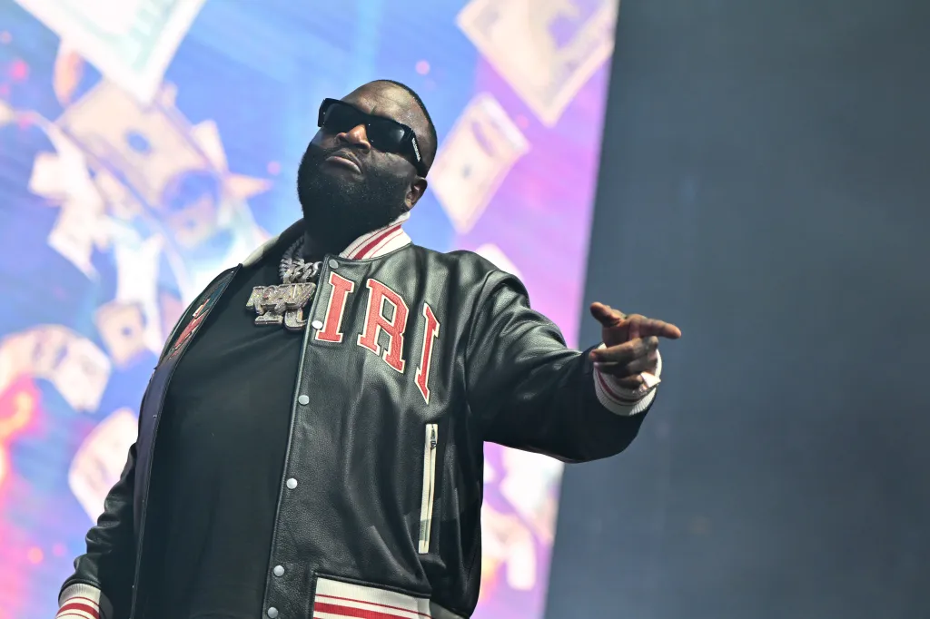 Rick Ross performs on stage, wearing a black bomber jacket with red lettering and dark sunglasses, with blurry dollar bills in the background.