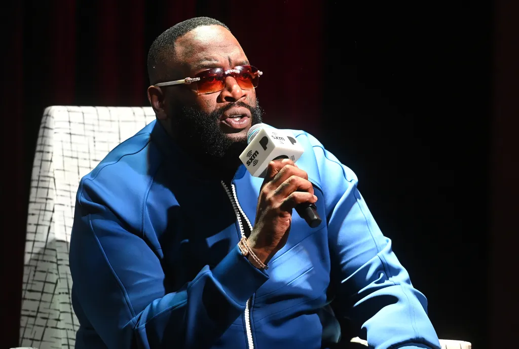 Rick Ross speaks onstage at Pandora Play Back, holding a microphone.