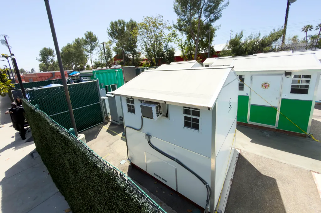 Homeless shelter with small white buildings, a green fence, and trees in the background.