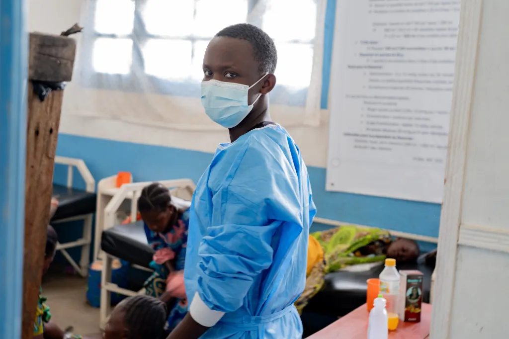 A health worker in a face mask and blue scrubs looks back at the camera inside a hospital ward.