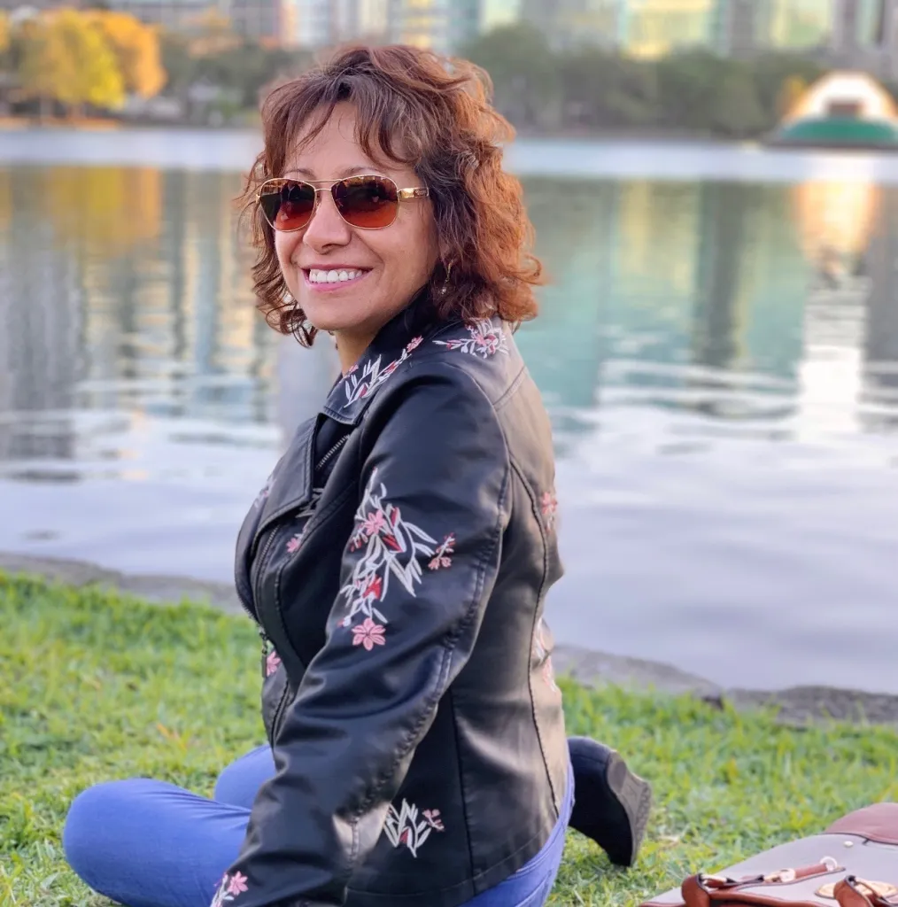 Rita Loncharich, a 65-year-old woman, smiling while sitting on grass next to a body of water.