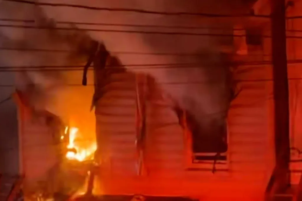 A house engulfed in large flames and smoke, with bright orange fire visible through windows and doors.