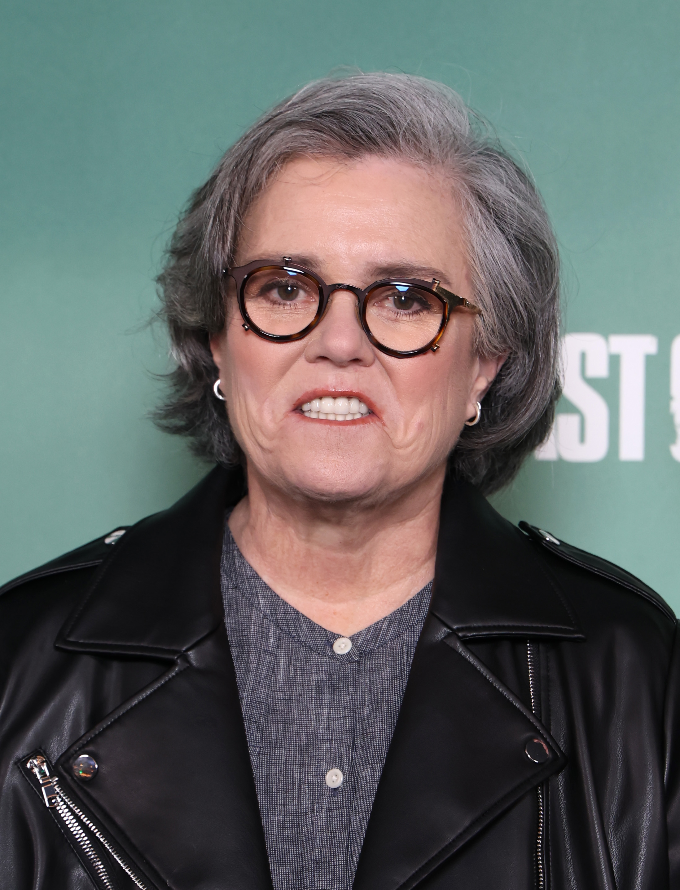 Rosie O'Donnell in a black leather jacket and glasses, smiling at a premiere.