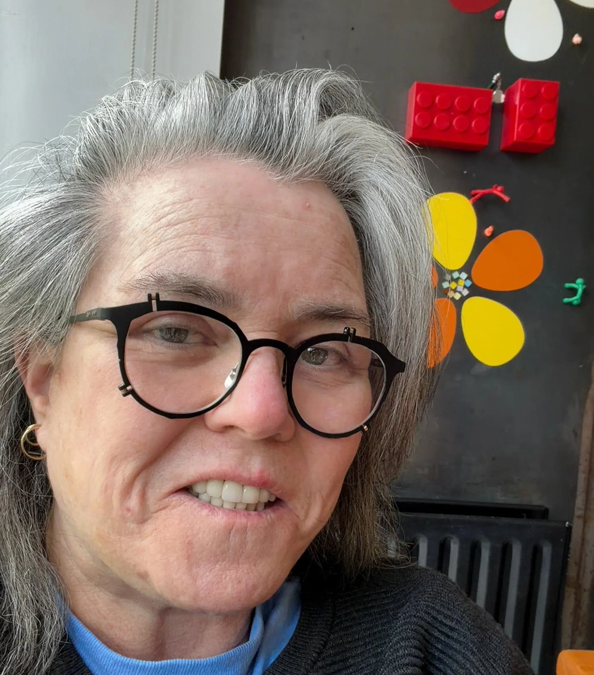 Rosie O'Donnell in a close-up selfie, wearing glasses and a light blue shirt.