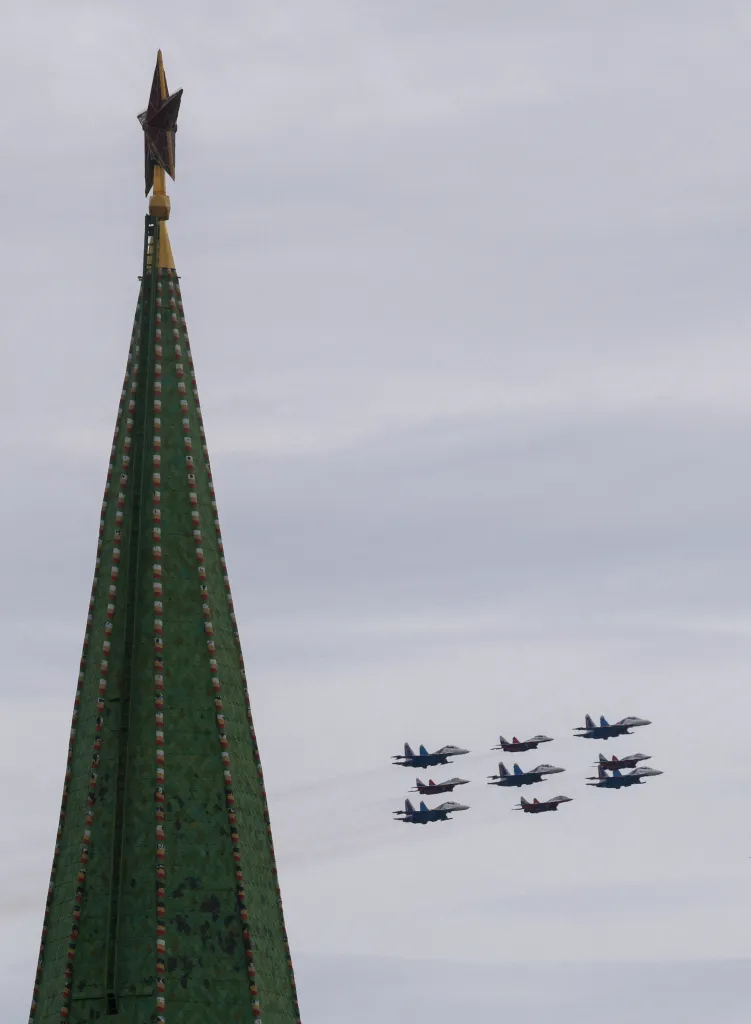 MiG-29 and Su-30SM jet fighters fly in formation over a green steeple with a red star on top during a military parade.