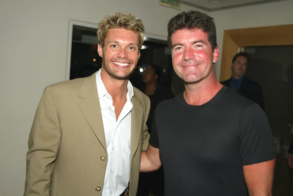 Ryan Seacrest in a tan suit and Simon Cowell in a black t-shirt smile at the camera.