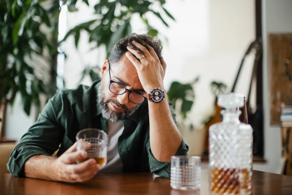 A sad man with a beard and glasses holds his head in his hand while drinking whiskey.