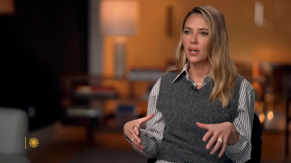Scarlett Johansson with long blonde hair, wearing a striped shirt and gray sweater vest, speaking and gesturing with her hands.