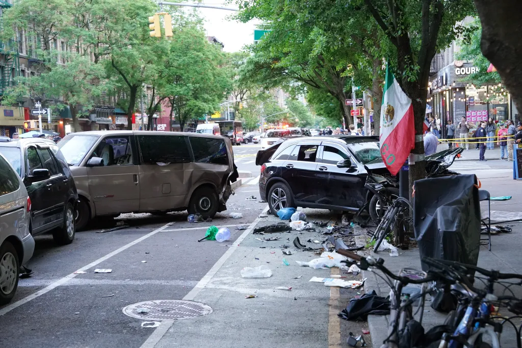 Two damaged cars, debris, and a crumpled bicycle litter a New York City sidewalk after an accident.