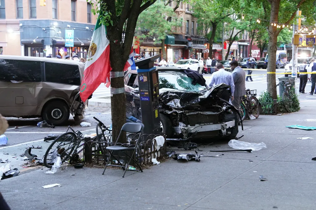 A black car with a smashed front end and a tan minivan with a damaged side, crashed on a New York City sidewalk, with debris scattered around.