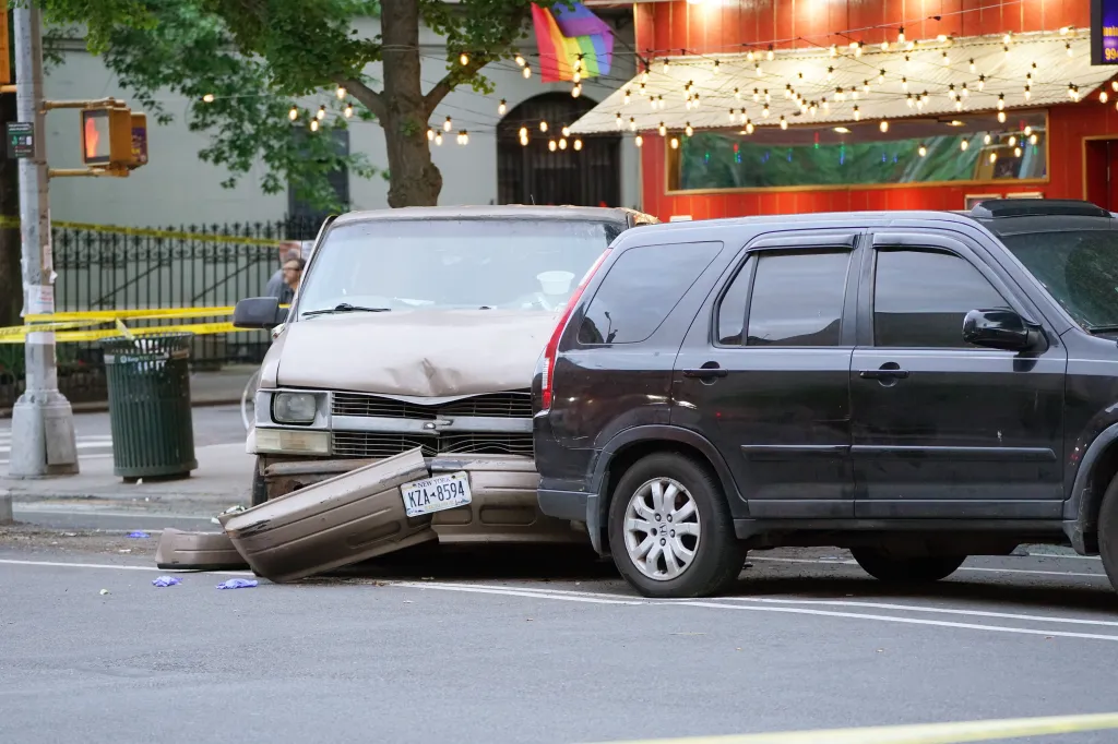 A damaged tan van with its bumper detached from a collision and a black SUV on a street cordoned off by yellow tape.