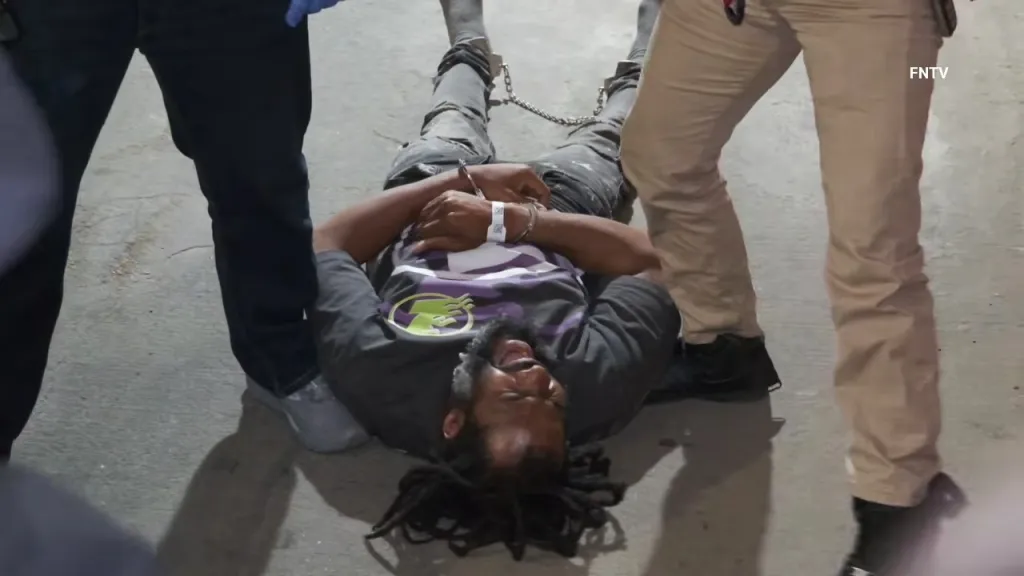 A man with dreadlocks lies on the ground, handcuffed at wrists and ankles, while two individuals stand over him.